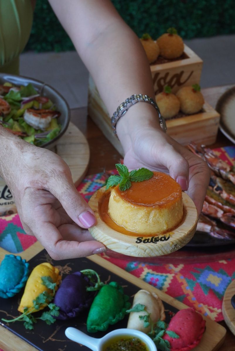 Hands lifting a Salsa leche flan above a colourful table of empanadas and shared plates.