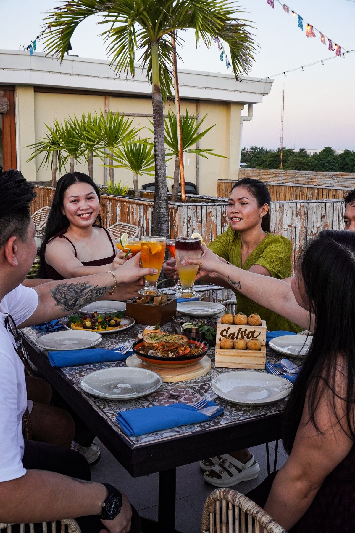 Friends toasting over dinner at a rooftop table at Salsa, beneath palm trees at golden hour.